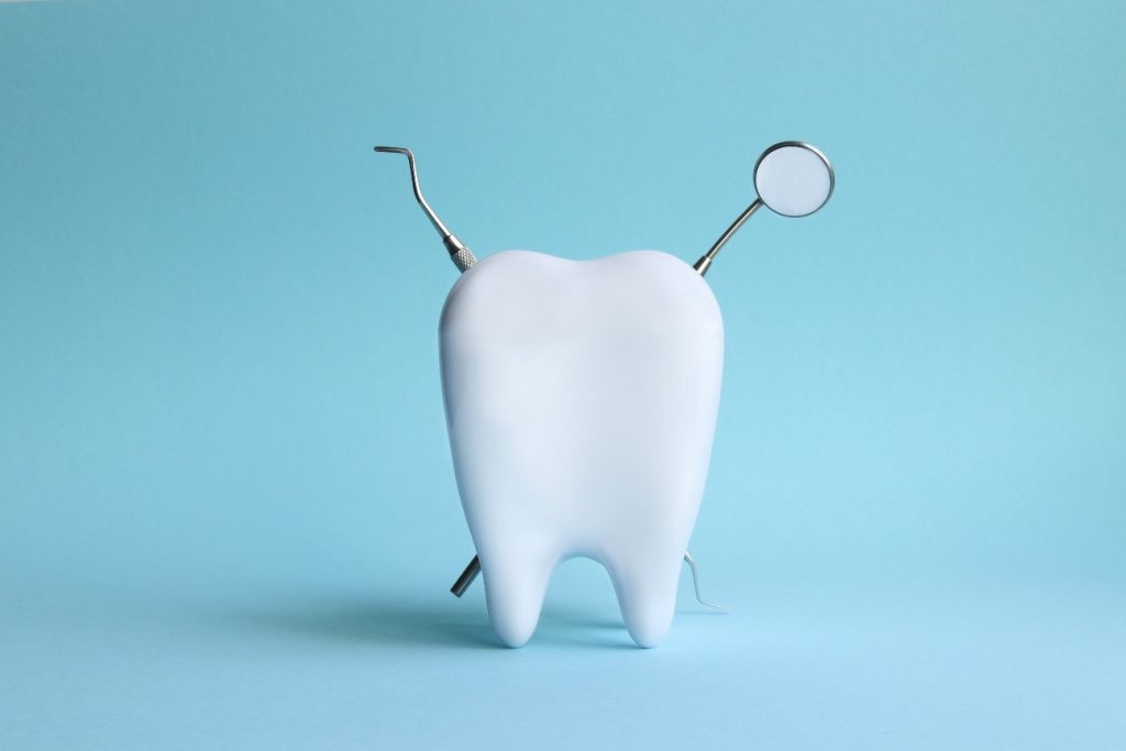 No centro da imagem de fundo azul claro há um dente, atrás do dente há dois instrumentos odontológicos: um espelho clínico e uma sonda exploradora, ambos formam um X.