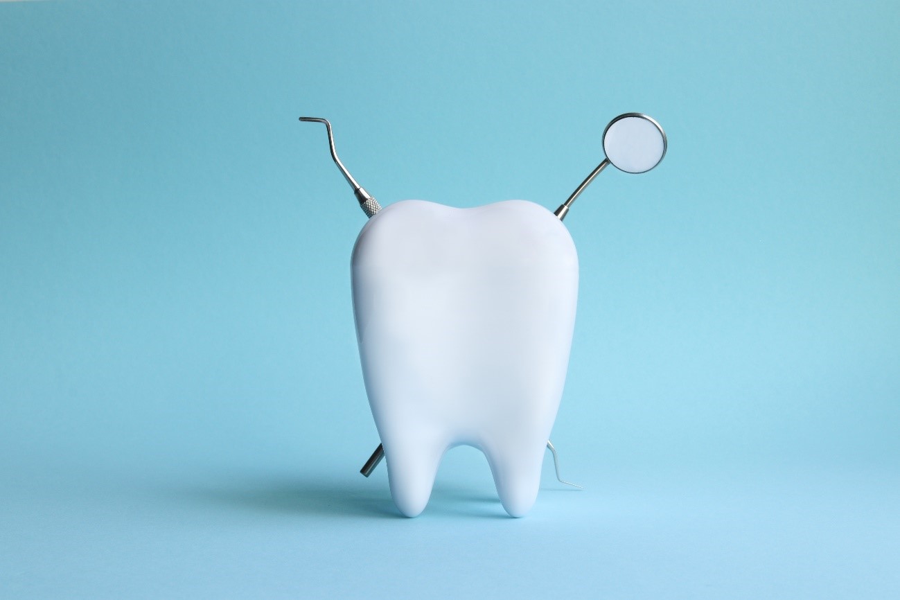 No centro da imagem de fundo azul claro há um dente, atrás do dente há dois instrumentos odontológicos: um espelho clínico e uma sonda exploradora, ambos formam um X.